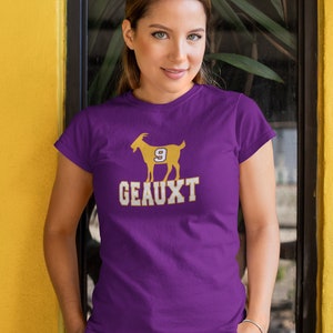 May include: Purple t-shirt with a gold silhouette of a goat wearing a jersey with the number 9. The text "GEAUX" is printed below the goat.