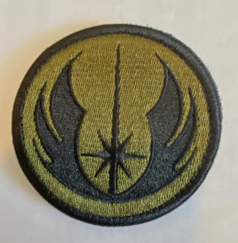 Peut inclure: Un &eacute;cusson brod&eacute; vert et noir avec un sabre laser stylis&eacute; et des ailes. L'&eacute;cusson est circulaire et a une bordure noire.