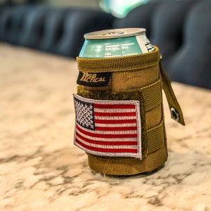 Puede incluir: Un portavasos t&aacute;ctico de color marr&oacute;n claro con un parche de velcro de la bandera estadounidense y el texto "75B Tactical" en la parte delantera. El portavasos contiene una lata de cerveza con el texto "ALCOHOL IBU 4.0% 17" en el lateral.