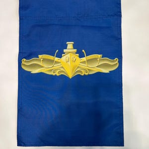 Puede incluir: Una bandera de jard&iacute;n azul con un emblema de submarino dorado. El emblema presenta un submarino con dos delfines flanqueantes y un tridente.