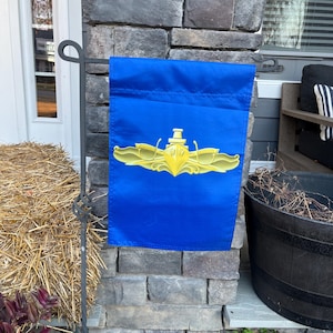 Puede incluir: Una bandera de jard&iacute;n azul con un emblema de submarino dorado. El emblema es un submarino estilizado con alas y una h&eacute;lice. La bandera est&aacute; colgada de un asta de bandera de metal negro.