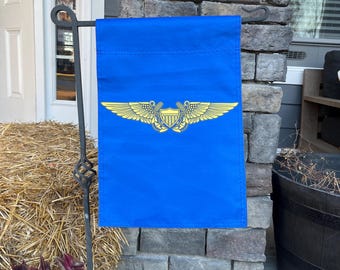 Puede incluir: Una bandera de jardín azul con un diseño bordado en oro de un par de alas con un escudo en el centro. El escudo tiene un diseño rojo, blanco y azul.