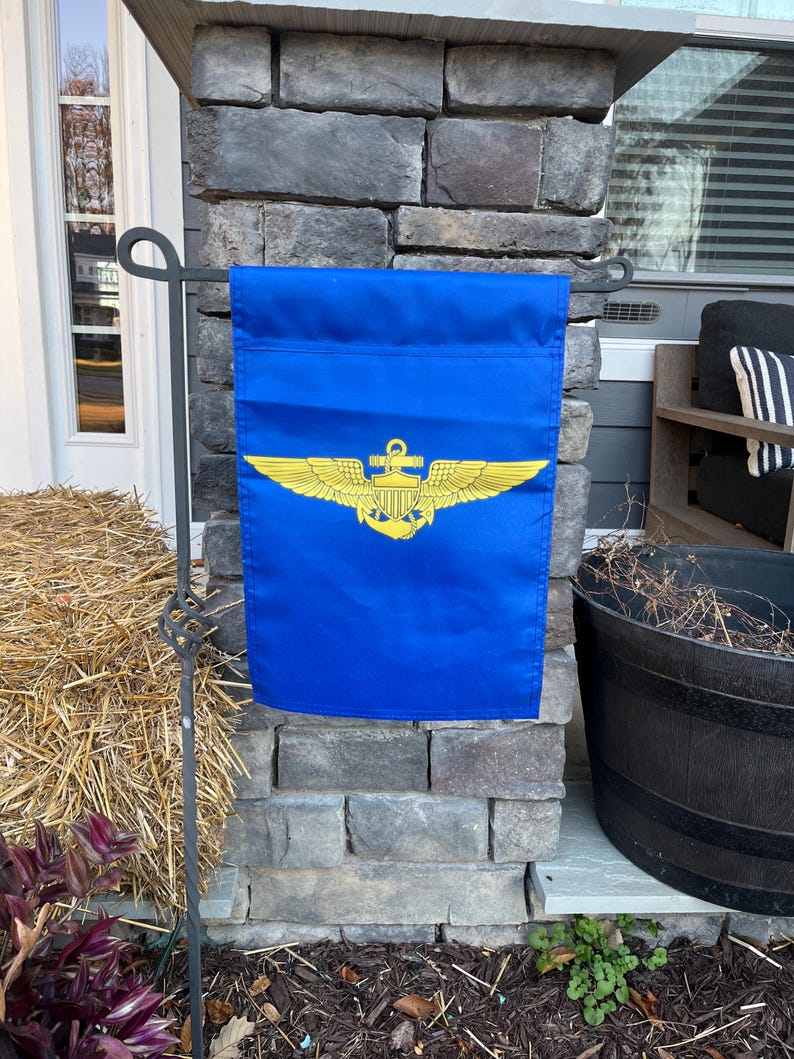 May include: A blue garden flag with a gold naval aviator wings and anchor insignia. The flag is hanging on a black metal flagpole.