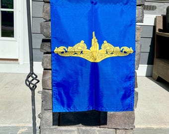 Puede incluir: Una bandera azul con un emblema dorado que presenta un diseño de submarino. La bandera es rectangular y cuelga verticalmente. El emblema incluye un submarino, peces y otros elementos náuticos. La bandera se muestra al aire libre.