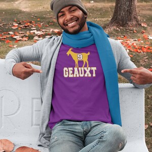 May include: A purple t-shirt with a yellow goat graphic and the text "GEAUX 9" printed on the front. The person wearing the shirt is smiling and pointing at the shirt.
