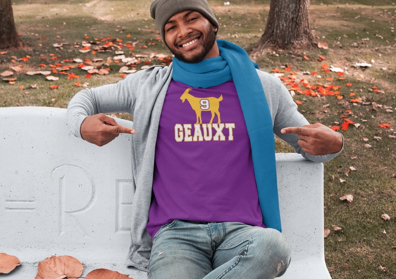 May include: A purple t-shirt with a yellow goat graphic and the text "GEAUX 9" printed on the front. The person wearing the shirt is smiling and pointing at the shirt.