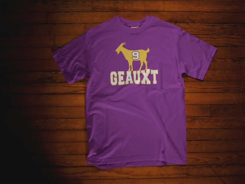 May include: Purple t-shirt with a gold silhouette of a goat wearing the number 9 and the word "GEAUX" printed on the front.