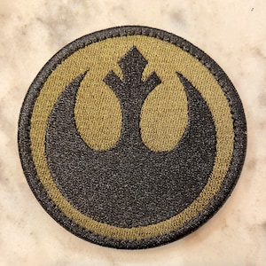 Peut inclure: Un &eacute;cusson rond brod&eacute; de couleur olive avec un symbole noir de l'Alliance rebelle. Le symbole est un oiseau stylis&eacute; avec les ailes d&eacute;ploy&eacute;es.