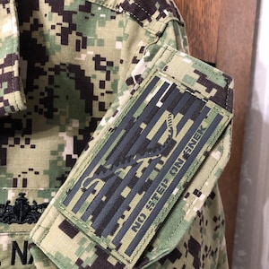 May include: A camouflage military uniform shirt with a black patch on the sleeve. The patch features a stylized American flag with the text "NO STEP ON SNAKE".