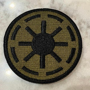 Peut inclure: Un &eacute;cusson brod&eacute; noir et vert avec le symbole de l'Empire galactique. Le symbole est un cercle avec une &eacute;toile noire au centre et quatre lignes noires s'&eacute;tendant de l'&eacute;toile jusqu'au bord du cercle.