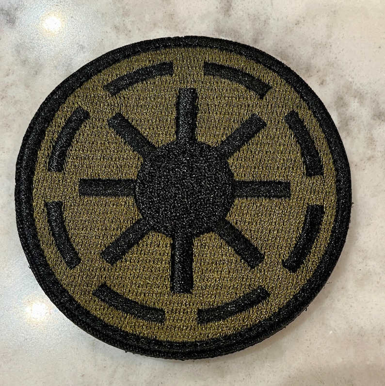 Peut inclure: Un &eacute;cusson brod&eacute; noir et vert avec le symbole de l'Empire galactique. Le symbole est un cercle avec une &eacute;toile noire au centre et quatre lignes noires s'&eacute;tendant de l'&eacute;toile jusqu'au bord du cercle.