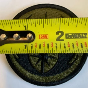 Peut inclure: Un patch brod&eacute; vert et noir avec un motif de boussole. Le patch est mesur&eacute; avec un ruban &agrave; mesurer jaune, montrant qu'il mesure environ 5 cm de diam&egrave;tre.
