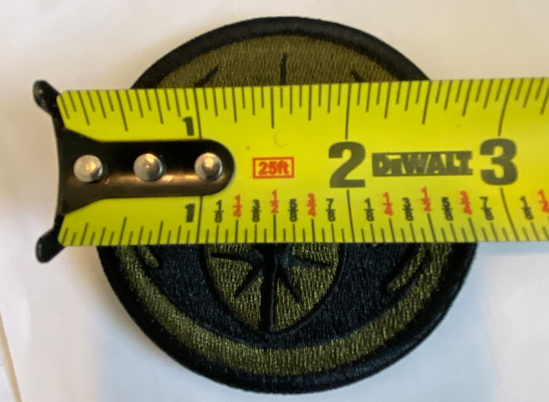 Peut inclure: Un patch brod&eacute; vert et noir avec un motif de boussole. Le patch est mesur&eacute; avec un ruban &agrave; mesurer jaune, montrant qu'il mesure environ 5 cm de diam&egrave;tre.