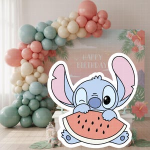 Może przedstawiać: Niebieska postać z kresk&oacute;wki, trzymająca kawałek arbuza, mruga przed tłem z napisem "Happy Birthday". Łuk balonowy w pastelowych kolorach i tropikalne kwiaty dopełniają świąteczną scenę.