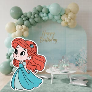 May include: A cartoon illustration of a mermaid in a blue dress with red hair and a blue flower. The background features a balloon arch in shades of green and cream, a backdrop with the words "Happy Birthday", and a small table with cupcakes.