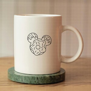 May include: A white ceramic mug featuring a black line art graphic of three donuts arranged in the shape of a mouse head silhouette. The mug has a smooth finish, a cylindrical body, and a curved handle on the right side, centered on a round base.