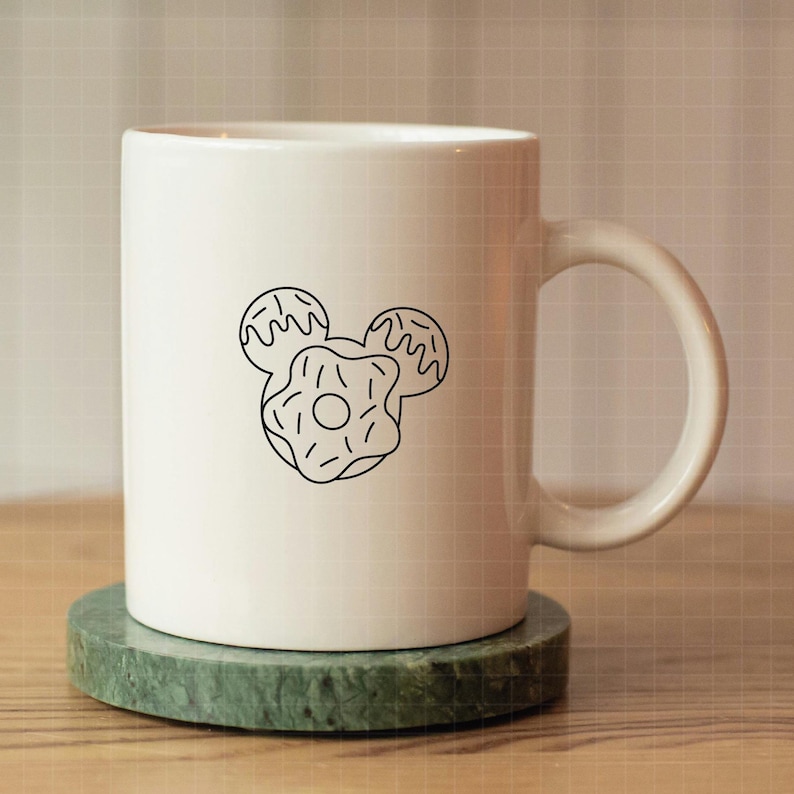 May include: A white ceramic mug featuring a black line art graphic of three donuts arranged in the shape of a mouse head silhouette. The mug has a smooth finish, a cylindrical body, and a curved handle on the right side, centered on a round base.
