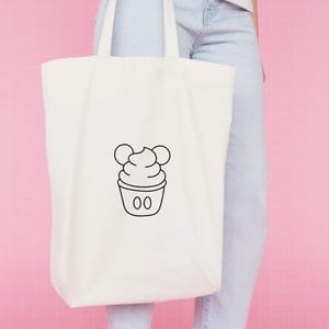May include: A white canvas tote bag featuring a centered black line art graphic of a soft serve ice cream cone with two circular ears at the top and two small vertical ovals on the base, resembling a classic character-inspired dessert design.