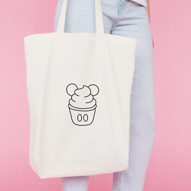 May include: A white canvas tote bag featuring a centered black line art graphic of a soft serve ice cream cone with two circular ears at the top and two small vertical ovals on the base, resembling a classic character-inspired dessert design.
