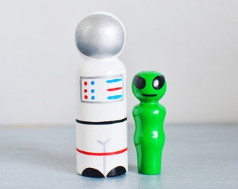 May include: A white astronaut peg doll with a silver helmet and a green alien peg doll. The astronaut has red and blue details. The alien has black eyes and a smile. The toys are standing side by side.