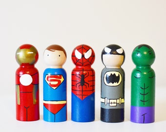 May include: Five wooden peg dolls painted as superheroes. The dolls are painted in red, blue, green, gray, and red and black. The dolls are standing on a white surface.