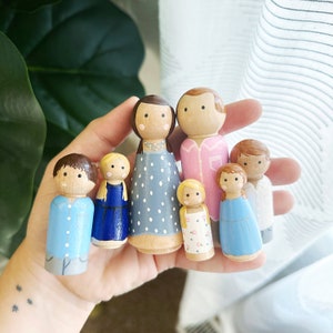 May include: A set of seven wooden peg dolls, painted in various colors and designs. The dolls are dressed in different outfits, including a blue shirt, a pink dress, a blue dress, a white dress with polka dots, and a floral dress. The dolls are arranged in a row, with the tallest doll in the center.