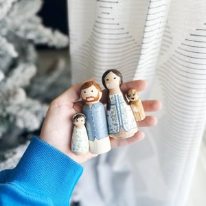May include: A set of four hand-painted wooden peg dolls, including a family of three and a dog. The dolls are dressed in blue and white clothing with floral patterns. The dog is brown and white.