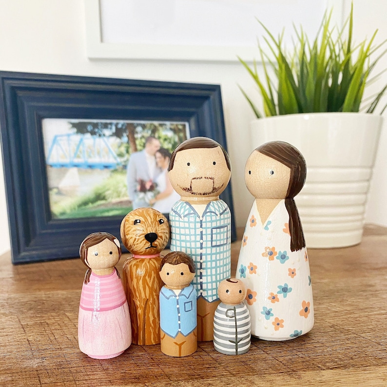 May include: A set of six wooden peg dolls, including a family of four and a dog. The dolls are painted in various colors, including blue, pink, green, and brown. The family members have painted facial features, and the dog has a brown and white coat.