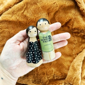 May include: Two wooden peg dolls, one wearing a black and white polka dot dress and the other wearing a green hooded sweatshirt with "THE NORTH FACE" printed on it.