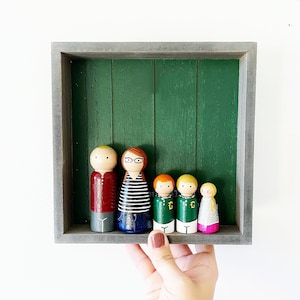 May include: A wooden frame with a green background holds a family of six wooden peg dolls. The dolls are painted in various colors and have different hairstyles and clothing. The dolls are arranged in a row, with the tallest doll in the center.