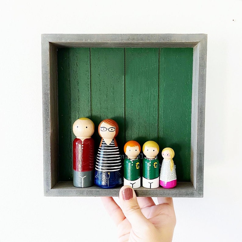 May include: A wooden frame with a green background holds a family of six wooden peg dolls. The dolls are painted in various colors and have different hairstyles and clothing. The dolls are arranged in a row, with the tallest doll in the center.