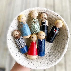 May include: A set of ten wooden peg dolls in a white crocheted basket. The dolls are painted in various colors and designs, including a blue and white striped shirt, a red dress, and a black and white striped shirt. The dolls are all wearing different expressions.