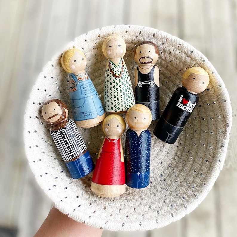 May include: A set of ten wooden peg dolls in a white crocheted basket. The dolls are painted in various colors and designs, including a blue and white striped shirt, a red dress, and a black and white striped shirt. The dolls are all wearing different expressions.