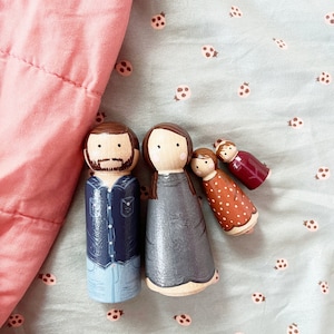 May include: A set of three wooden peg dolls, painted to resemble a family. The father doll is wearing a blue shirt and jeans, the mother doll is wearing a gray dress, and the child doll is wearing a red and orange dress with white polka dots.