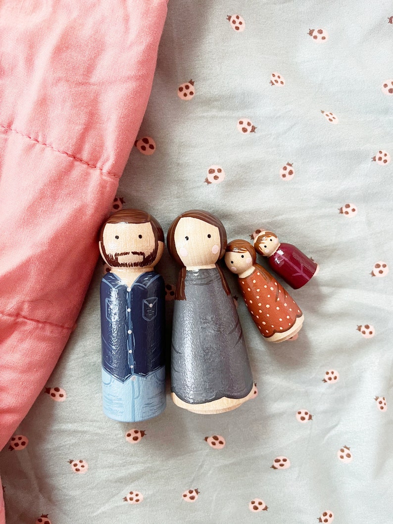 May include: A set of three wooden peg dolls, painted to resemble a family. The father doll is wearing a blue shirt and jeans, the mother doll is wearing a gray dress, and the child doll is wearing a red and orange dress with white polka dots.