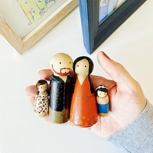 May include: A set of four wooden peg dolls, painted in a whimsical style. The dolls are a family, with a father, mother, and two children. The father has a brown beard and black hair, the mother has brown hair, and the children have red and brown hair. The dolls are dressed in colorful clothing.