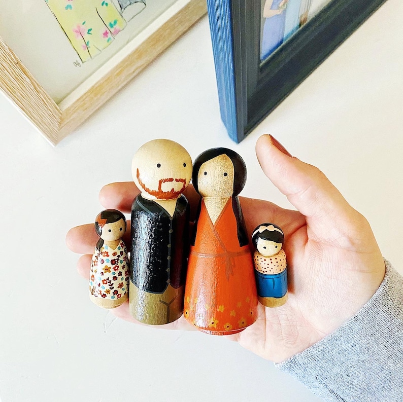 May include: A set of four wooden peg dolls, painted in a whimsical style. The dolls are a family, with a father, mother, and two children. The father has a brown beard and black hair, the mother has brown hair, and the children have red and brown hair. The dolls are dressed in colorful clothing.