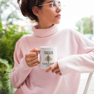 May include: White ceramic mug featuring the text "Sokka's Cactus Juice" with a desert landscape illustration. The mug has a handle and is held by a person wearing a pink turtleneck sweater.