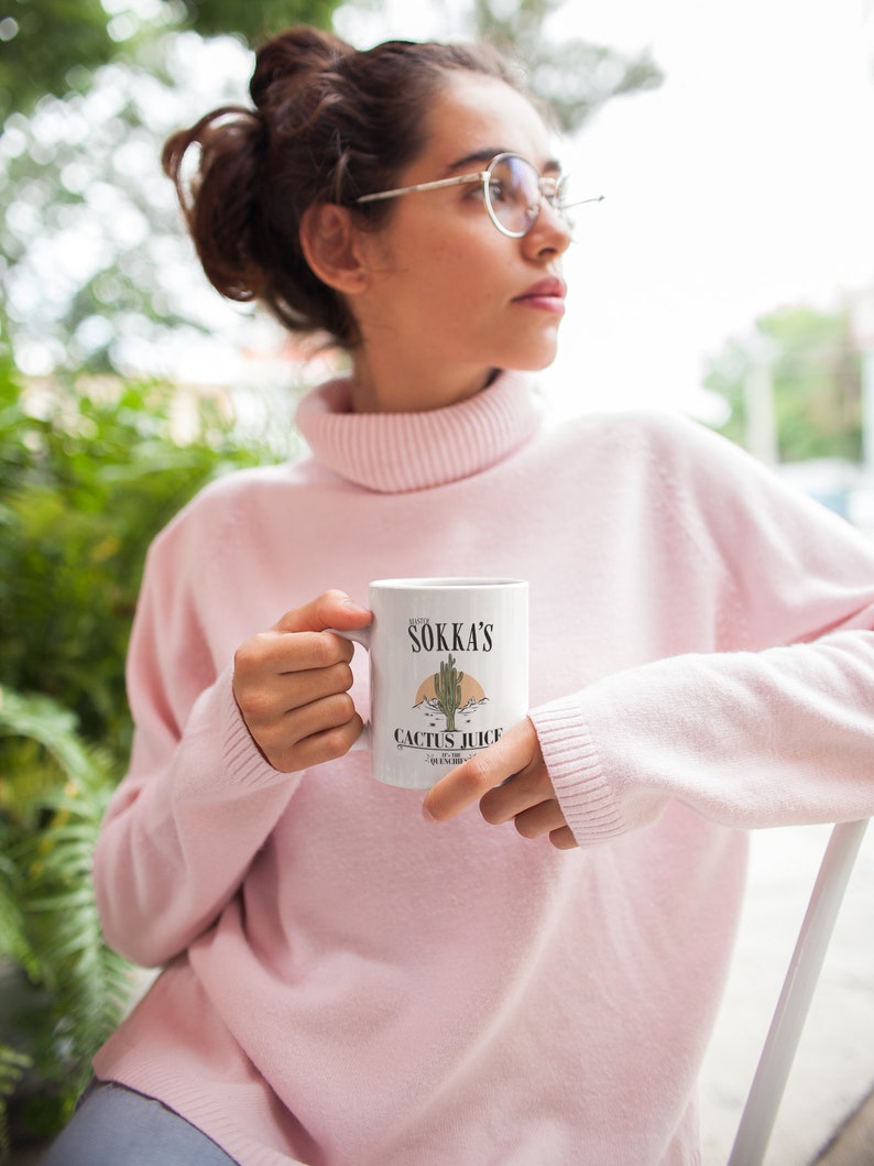 May include: White ceramic mug featuring the text "Sokka's Cactus Juice" with a desert landscape illustration. The mug has a handle and is held by a person wearing a pink turtleneck sweater.