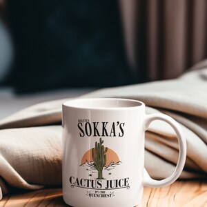 May include: White ceramic mug with a handle, featuring the text "Master Sokka's Cactus Juice" with a desert landscape illustration. The mug is on a wooden surface, with a blurred background.