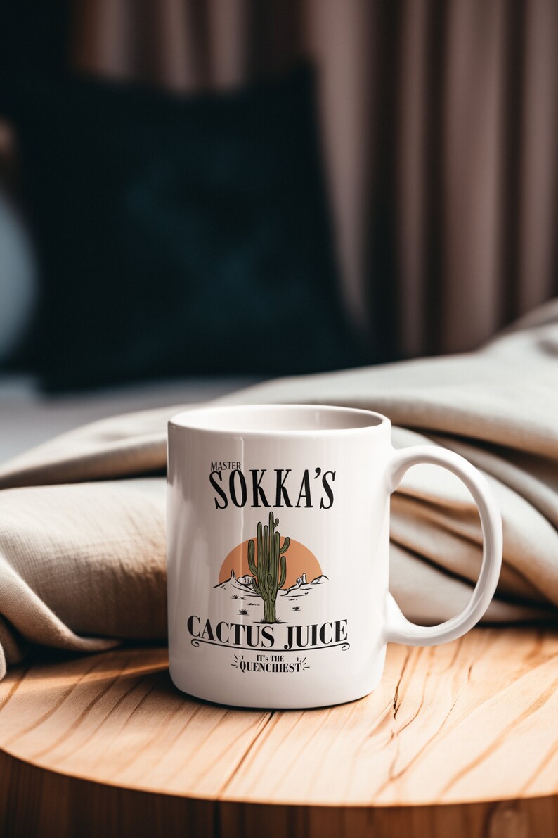 May include: White ceramic mug with a handle, featuring the text "Master Sokka's Cactus Juice" with a desert landscape illustration. The mug is on a wooden surface, with a blurred background.