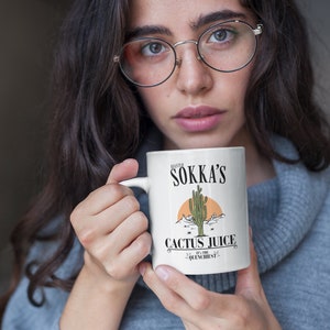 May include: White ceramic mug featuring the text "Master Sokka's Cactus Juice" with a desert landscape illustration. The mug has a handle and is held by a person. The text "It's the quenchiest!" is also on the mug.