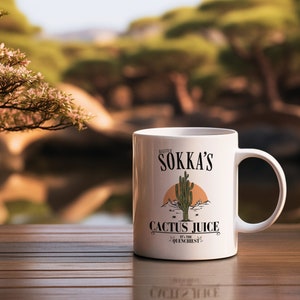 May include: White ceramic mug featuring the text "Master Sokka's Cactus Juice" with a desert landscape illustration. The mug has a handle and is set on a wooden surface with a blurred outdoor background.