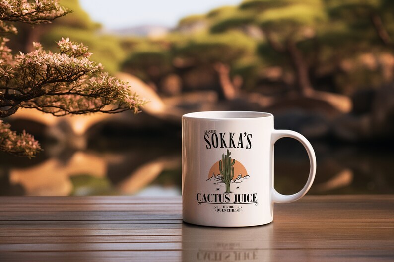 May include: White ceramic mug featuring the text "Master Sokka's Cactus Juice" with a desert landscape illustration. The mug has a handle and is set on a wooden surface with a blurred outdoor background.