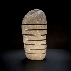May include: A carved stone sculpture with horizontal cut-out sections. The sculpture is a light gray color and has a rough, textured surface. The cuts create a layered effect, and the sculpture stands on a dark surface.