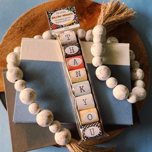 May include: A set of colorful wrapped candies with the word "THANK YOU" spelled out on them. The candies are on a wooden surface with a white beaded garland.