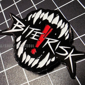 May include: Black enamel pin with the words "BITE RISK" in white, with a red exclamation point. The pin features a design of sharp white teeth and pointed edges. The pin is on a black and white grid.