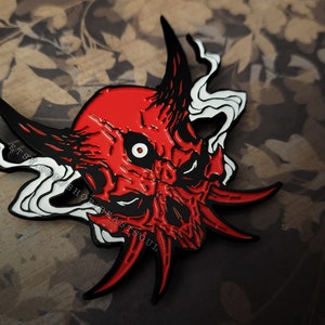 May include: Red and black enamel pin with a detailed skull design. The skull has a single white eye and sharp teeth. The pin is surrounded by white smoke-like designs.