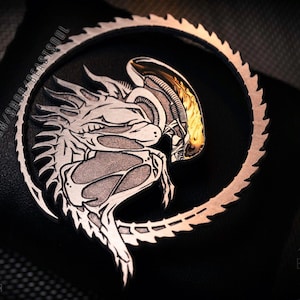 Op de afbeelding: Een zilveren en gouden metalen pin met een gedetailleerde illustratie van een buitenaards wezen met een cirkelvormig zaagbladontwerp. De pin is op een zwarte lederen achtergrond.