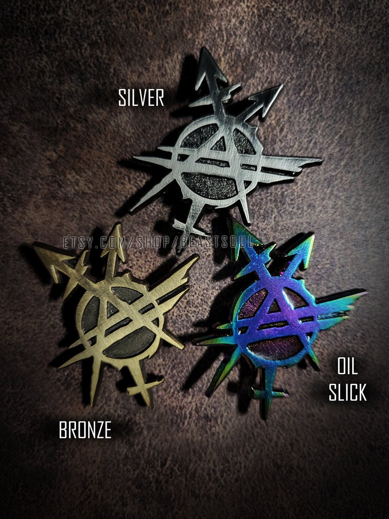 K&ouml;nnte beinhalten: Drei Metallanh&auml;nger in Silber-, Bronze- und &Ouml;leffekt-Ausf&uuml;hrung. Jeder Anh&auml;nger hat ein zentrales Symbol mit strahlenf&ouml;rmigen Pfeilen und einem stilisierten Buchstaben "A". Die W&ouml;rter "SILVER", "BRONZE" und "OIL SLICK" sind auf dem Bild aufgedruckt.